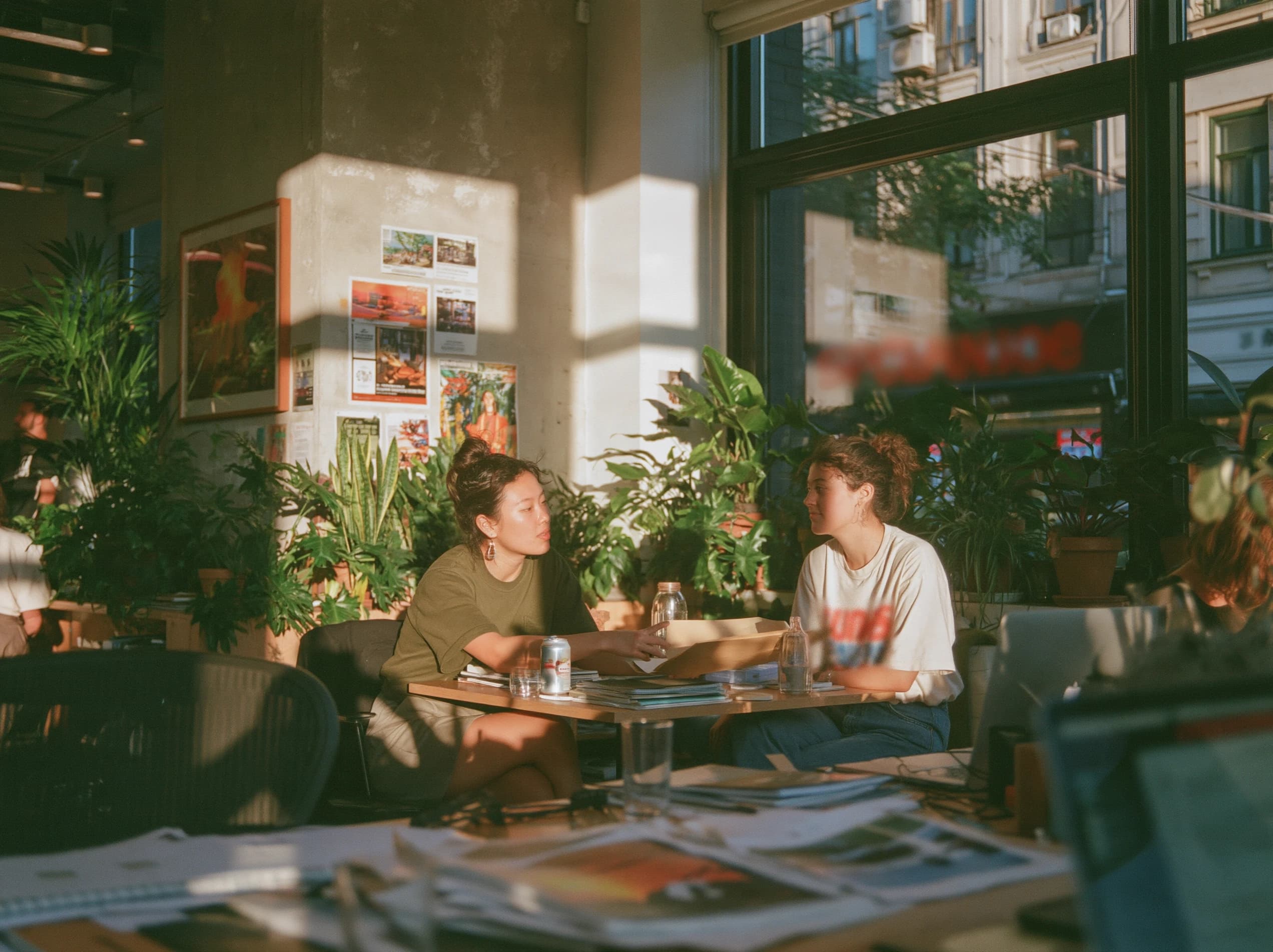 Zwei Frauen an einem kleinen Tisch in einem sonnigen Büro voller Pflanzen, die eine schiebt der anderen eine Mappe zu.