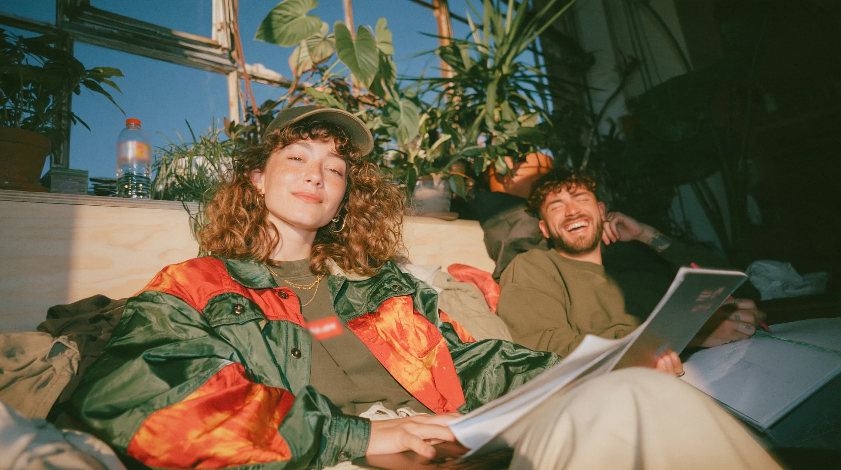 Two colleagues sitting relaxed by a sunny window full of plants, she smiles into the camera and he laughs over his notes.