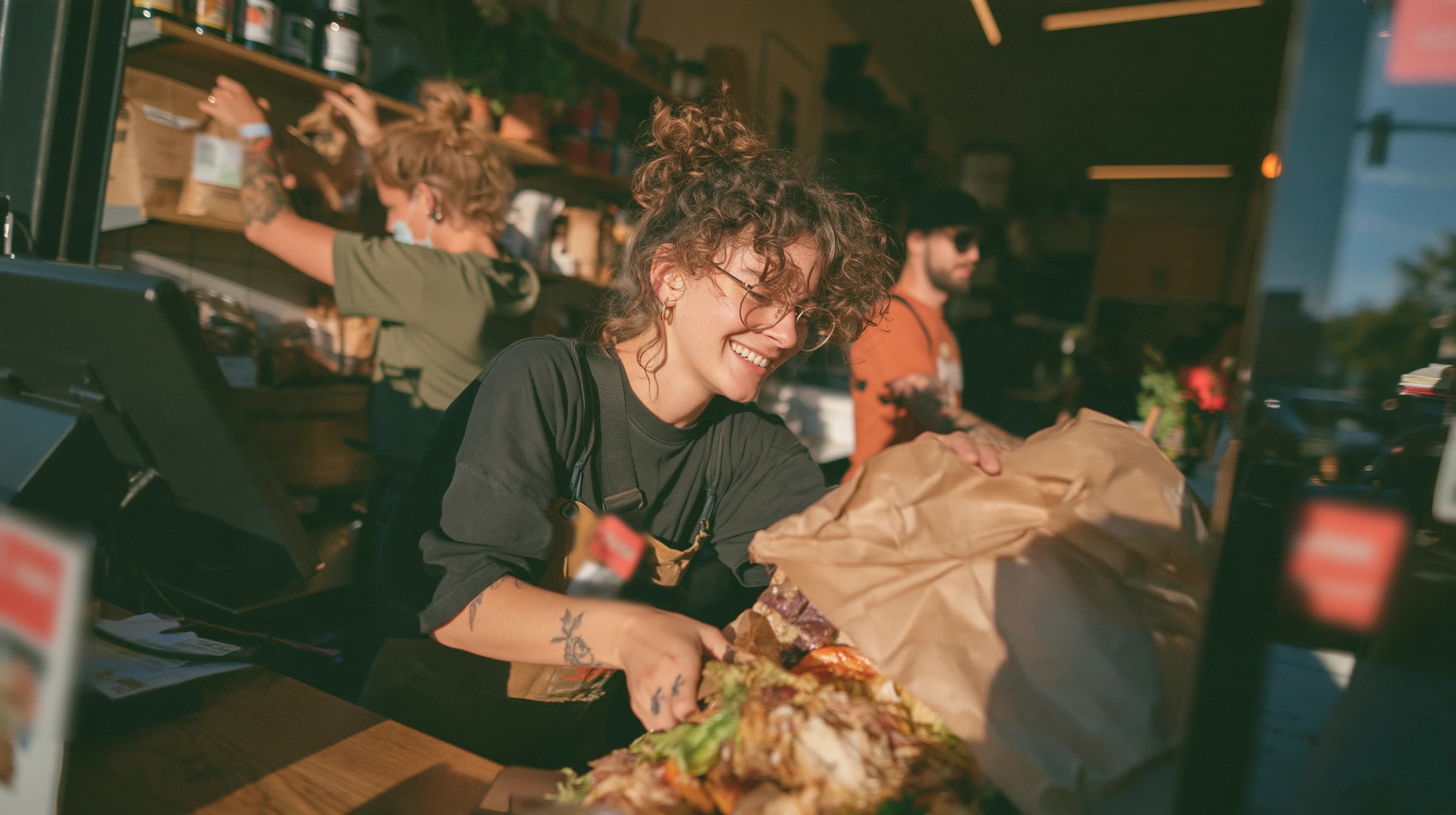 A smiling shop assistant packs food into a paper bag at the counter while her colleagues work behind her.
